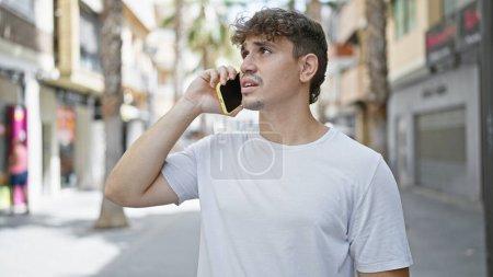 Cool aussehender, gut aussehender junger hispanischer Mann, der sich ernsthaft mit seinem Smartphone unterhält und mit konzentriertem Gesichtsausdruck vor der lässigen Modekulisse der sonnigen urbanen Straße steht.