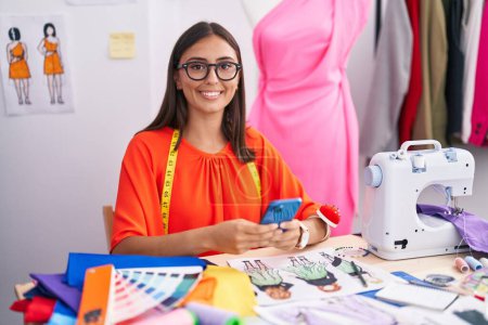 Téléchargez les photos : Jeune belle hispanique femme tailleur souriant confiant en utilisant smartphone au magasin de tailleur - en image libre de droit