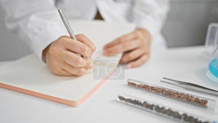 Photo for Dedicated hispanic woman scientist engrossed in study, scribbling notes in lab notebook, indoor scene expressing passion and work in biomedical science research - Royalty Free Image