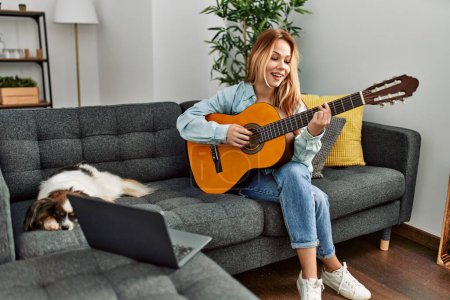 Foto de Joven mujer caucásica que tiene clase de guitarra clásica en línea sentado en el sofá con el perro en casa - Imagen libre de derechos