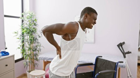 Téléchargez les photos : Homme afro-américain souffrant de maux de dos dans une salle de clinique de réadaptation avec équipement médical visible. - en image libre de droit