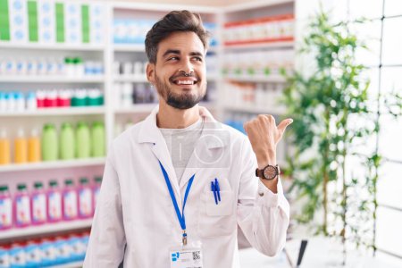 Foto de Joven hombre hispano con tatuajes trabajando en farmacia sonriendo con la cara feliz mirando y señalando a un lado con el pulgar hacia arriba. - Imagen libre de derechos