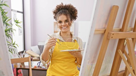 Photo for A smiling young hispanic woman with curly hair paints on a canvas in a bright art studio. - Royalty Free Image