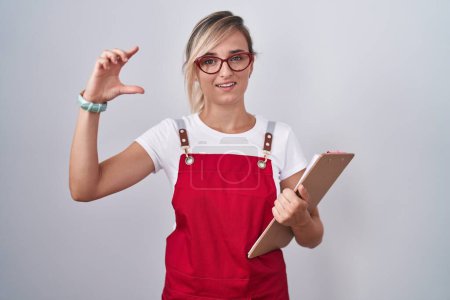 Photo for Young blonde woman wearing waiter uniform holding clipboard smiling and confident gesturing with hand doing small size sign with fingers looking and the camera. measure concept. - Royalty Free Image