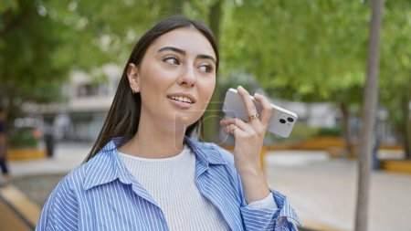 Photo for A young hispanic woman listens to a voicemail outdoors in a city park, showcasing a casual style and natural beauty. - Royalty Free Image