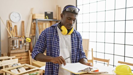 Téléchargez les photos : Homme afro-américain travaillant attentivement dans un atelier de menuiserie avec des outils, du bois et des équipements de sécurité. - en image libre de droit