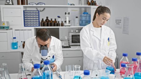 Photo for Hispanic man and woman scientists working together, engrossed in medical research at their indoor laboratory, measuring liquid samples while peering through their microscope. - Royalty Free Image