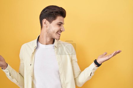 Téléchargez les photos : Jeune homme hispanique debout sur fond jaune souriant montrant les paumes ouvertes des deux mains, présentant et comparant la publicité et l'équilibre - en image libre de droit