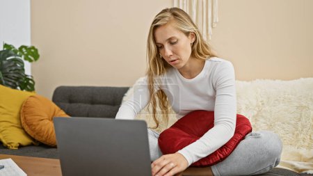 Téléchargez les photos : Une femme blonde concentrée utilise un ordinateur portable dans un salon confortable, incarnant le travail du confort et de la concentration à la maison. - en image libre de droit