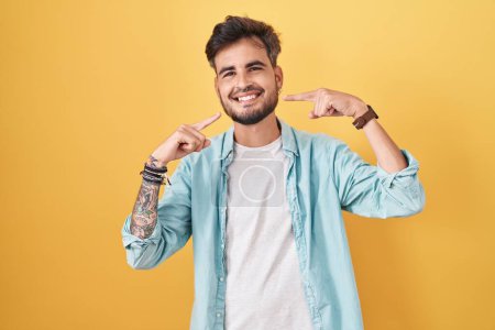 Téléchargez les photos : Young hispanic man with tattoos standing over yellow background smiling cheerful showing and pointing with fingers teeth and mouth. dental health concept. - en image libre de droit