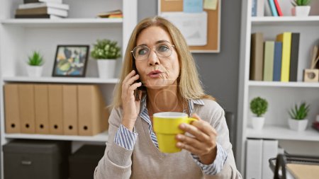 Foto de Una mujer madura bebe café mientras habla por teléfono en su oficina moderna, lo que indica una vida profesional ocupada. - Imagen libre de derechos
