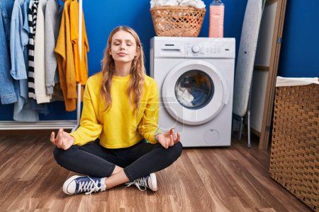 Téléchargez les photos : Young blonde woman doing yoga exercise waiting for washing machine at laundry room - en image libre de droit