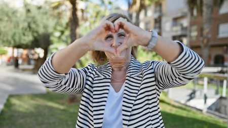 Téléchargez les photos : Femme mûre souriante faisant forme de coeur avec les mains dans le parc ensoleillé - en image libre de droit