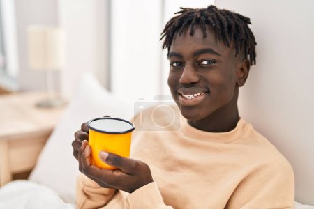 Foto de African american man drinking cup of coffee sitting on bed at bedroom - Imagen libre de derechos