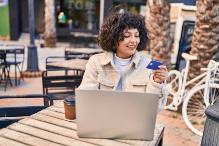 Foto de Joven mujer hispana hermosa usando portátil y tarjeta de crédito sentado en la mesa en la terraza de la cafetería - Imagen libre de derechos