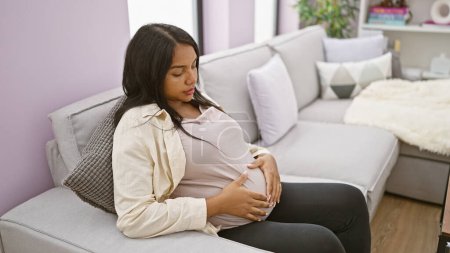 Foto de Mujer joven embarazada de cara seria sentada en casa, tocando casualmente su vientre en el sofá de su sala de estar, encarnando la emoción y la anticipación relajada de la maternidad. - Imagen libre de derechos