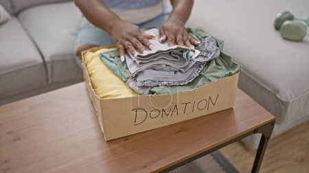 Téléchargez les photos : Femme afro-américaine chaleureuse mettant des vêtements pliés dans une boîte en carton pour un don de charité, confortablement assise sur son canapé du salon, transformant les corvées en soutien à la maison - en image libre de droit