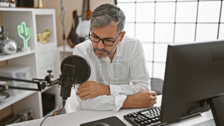 Foto de Un joven hispano de cabello gris al aire en el estudio de radio, un periodista serio con barba, gafas y mirada concentrada, hablando en un programa de radiodifusión profesional - Imagen libre de derechos