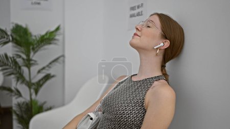 Téléchargez les photos : Jeune femme blonde assise sur une chaise écoutant de la musique détendue dans la salle d'attente - en image libre de droit