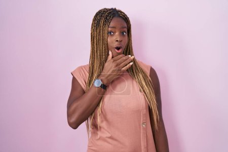 Téléchargez les photos : Femme afro-américaine avec des cheveux tressés debout sur fond rose, fascinée par l'incrédulité, la surprise et l'expression étonnante avec les mains sur le menton - en image libre de droit