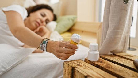 Téléchargez les photos : Femme hispanique couché dans le lit tenant des médicaments dans une scène de chambre confortable - en image libre de droit