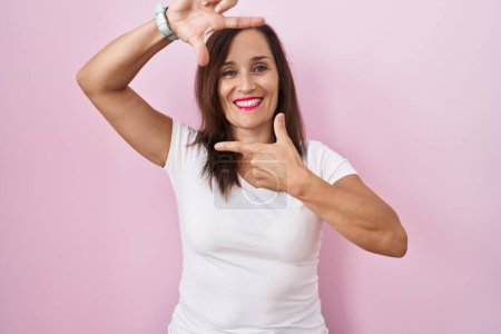 Téléchargez les photos : Femme brune du moyen âge debout sur fond rose souriant faisant cadre avec les mains et les doigts avec le visage heureux. concept de créativité et photographie. - en image libre de droit