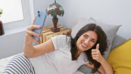 Foto de Mujer sonriente usando un teléfono inteligente para una selfie en un acogedor dormitorio, que representa la relajación casual y la felicidad. - Imagen libre de derechos