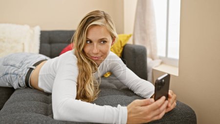 Téléchargez les photos : Une jeune femme blonde séduisante en tenue décontractée prend un selfie sur un canapé dans un intérieur confortable de la maison. - en image libre de droit
