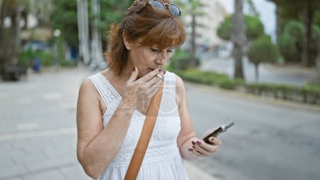 Foto de Una pelirroja madura fuma y comprueba su teléfono en una calle urbana soleada. - Imagen libre de derechos