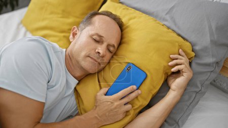 Photo for Exhausted middle age man cozily ensconced in comfort of his bedroom, resting on bed with smartphone, surrendering to relaxing pleasure of sleep - Royalty Free Image