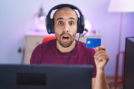 Foto de Hombre hispano con barba haciendo compras en línea con computadora y tarjeta de crédito asustado y sorprendido con la boca abierta para sorpresa, cara de incredulidad - Imagen libre de derechos