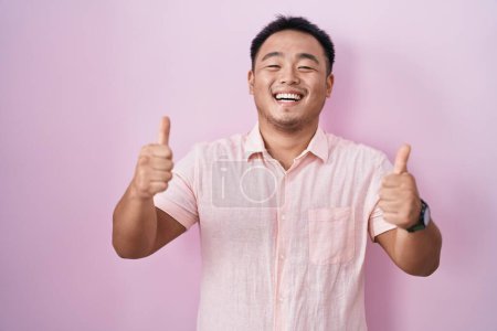 Foto de Joven chino de pie sobre el signo de éxito de fondo rosa haciendo gesto positivo con la mano, pulgares hacia arriba sonriendo y feliz. expresión alegre y gesto ganador. - Imagen libre de derechos