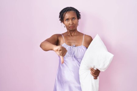Foto de Mujer africana con rastas usando pijama abrazando la almohada luciendo infeliz y enojada mostrando rechazo y negativa con el gesto de los pulgares hacia abajo. mala expresión. - Imagen libre de derechos