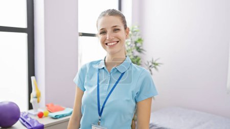 Téléchargez les photos : Portrait d'une jeune femme souriante portant une chemise bleue et un insigne, debout dans une salle de clinique de réadaptation lumineuse. - en image libre de droit