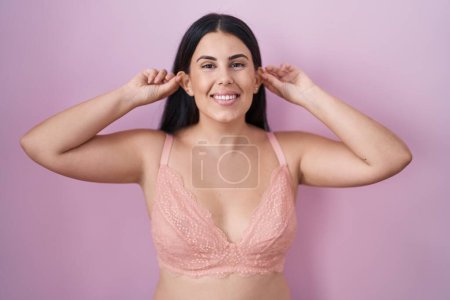 Foto de Mujer hispana joven con sujetador rosa sonriendo tirando de las orejas con los dedos, gesto divertido. problema de audición - Imagen libre de derechos