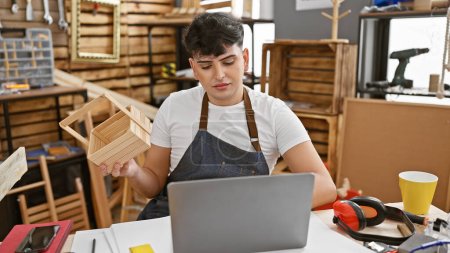 Photo for Man examines handmade wooden shelf in a well-equipped carpentry workshop while using laptop - Royalty Free Image