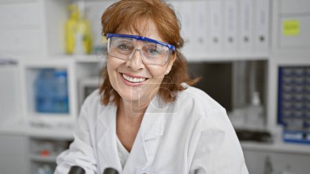 Foto de Científica madura sonriente en bata de laboratorio y gafas protectoras en entorno de laboratorio - Imagen libre de derechos