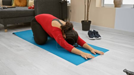 Foto de Una joven se estira sobre una esterilla de yoga azul en una sala de estar bien iluminada, con ropa deportiva y rodeada de una decoración minimalista. - Imagen libre de derechos