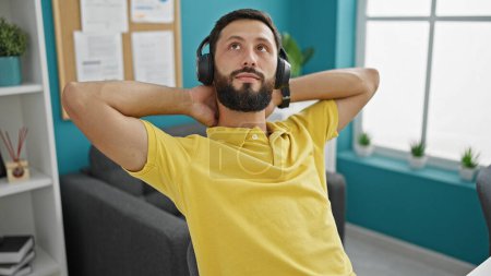 Foto de Joven trabajador de negocios hispano escuchando música relajado con las manos en la cabeza en la oficina - Imagen libre de derechos