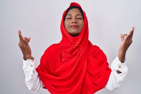 Foto de Mujer árabe joven vistiendo bufanda islámica tradicional hijab relajado y sonriendo con los ojos cerrados haciendo gesto de meditación con los dedos. concepto de yoga. - Imagen libre de derechos