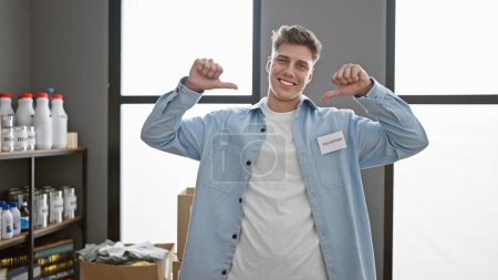 Photo for Young, confident, and smiling, a caucasian man volunteer points to himself at the charity center. surrounded by donation boxes, his altruistic spirit stands out bright in the warehouse. - Royalty Free Image