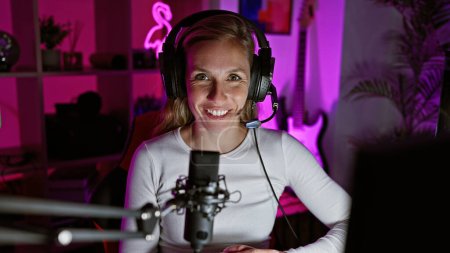 Foto de Mujer sonriente con auriculares en una sala de juegos iluminada por neón con micrófono y computadora por la noche. - Imagen libre de derechos