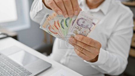 Foto de Un hombre de mediana edad en una oficina tiene moneda singapurense, retratando las finanzas, los negocios, la computadora portátil y el profesionalismo. - Imagen libre de derechos