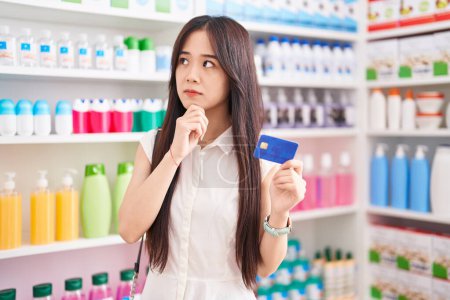 Photo for Young chinese woman shopping at pharmacy drugstore holding credit card serious face thinking about question with hand on chin, thoughtful about confusing idea - Royalty Free Image