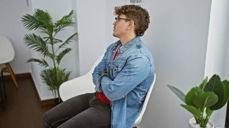 Téléchargez les photos : Beau jeune homme hispanique à l'intérieur, sérieusement concentré, assis dans la chaise du hall avec les bras croisés dans la salle d'attente - en image libre de droit