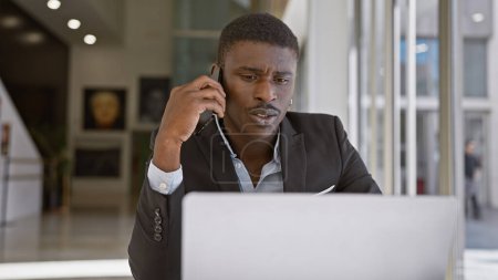 Foto de Hombre africano de traje trabajando en el ordenador portátil y hablando por teléfono en la oficina moderna - Imagen libre de derechos