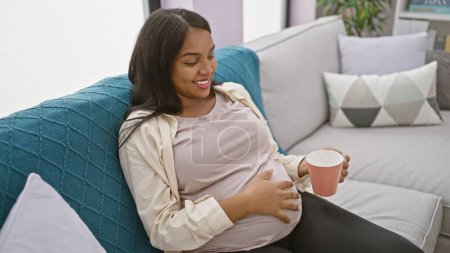 Foto de Mujer joven radiante, esperándola primero, disfruta de una mañana relajante en casa, cómodamente sentada en el sofá, sosteniendo una taza de café, tocando tiernamente su vientre embarazada mientras sonríe - Imagen libre de derechos