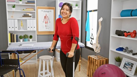 Photo for A smiling young hispanic woman using crutches in a well-equipped physical therapy room. - Royalty Free Image