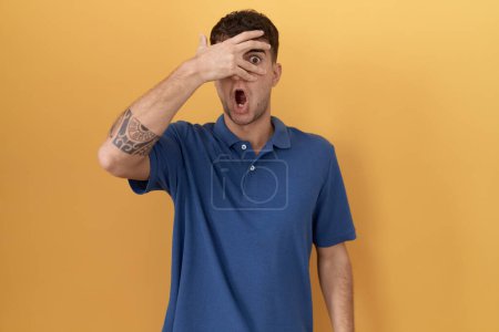 Foto de Young hispanic man standing over yellow background peeking in shock covering face and eyes with hand, looking through fingers with embarrassed expression. - Imagen libre de derechos