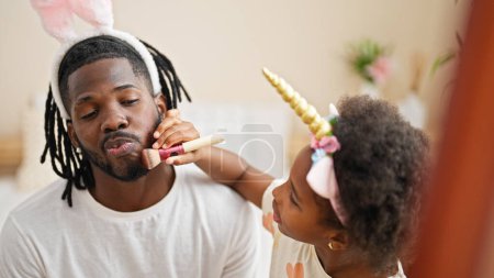 Foto de Padre e hija afroamericanos usando diadema divertida jugando con maquillaje en el dormitorio - Imagen libre de derechos